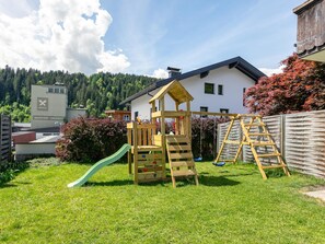 A wooden play structure with a slide and climbing ladder in a grassy yard, a white building with a dark roof in the background, and a forested hill.