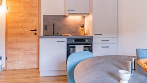 Une cuisine de taille réduite avec des meubles blancs, une porte en bois, un four encastré et une table ronde accompagnée d’une chaise bleue.