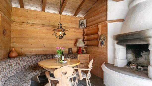 Un intérieur en bois avec un sol en pierre, une table ronde en bois entourée de chaises, un banc, une cheminée et un luminaire suspendu.