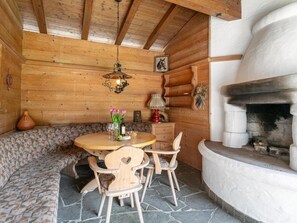 Een houten interieur met een stenen vloer, een ronde houten tafel met stoelen, een bank, een open haard en een hanglamp.