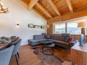 A modern living room with a wooden ceiling, a large window with a view, a grey sectional sofa, and a dining area with chairs and a table.