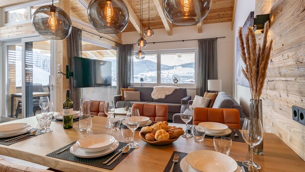 Un coin repas avec une table en bois dressée pour le repas, un canapé en arrière-plan et une vue sur des montagnes enneigées par de grandes fenêtres.