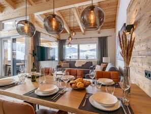 A dining area with a wooden table set for a meal, a sofa in the background, and a view of snowy mountains through large windows.