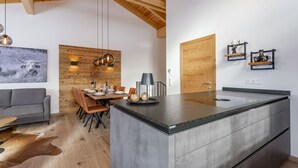 Une cuisine moderne avec un îlot central, des poutres en bois au plafond, un coin repas avec un mur en bois et un canapé gris.