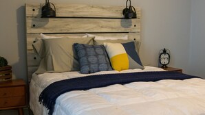 A neatly made bed with a wooden headboard, multiple pillows, a dark blue throw blanket, and a small bedside table with a clock. - Cozy Cottage within walking distance of downtown! (Bristol)