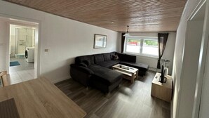 En moderne stue med træloft, en sort sofa, et sofabord og udsigt over et boligområde gennem et vindue.