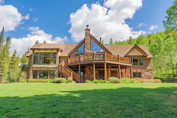 A large, two-story house with a prominent stone chimney, a wooden deck, and a large front lawn.
