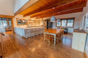 A spacious kitchen with a large island, wooden dining table, and chairs. There are multiple windows providing natural light.