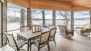 Ein überdachter Außenbereich mit Tisch und Stühlen, großen Fenstern und Blick auf ein Gewässer.