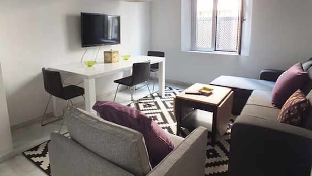 A modern living room with a white sofa, a small table, a TV mounted on the wall, and two pendant lights.