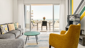 Ein modernes Wohnzimmer mit einer Couch, einem gelben Sessel, einem kleinen Tisch und einem Fernseher. Durch die Glastüren ist ein Balkon mit Blick auf die Stadt und das Meer zu sehen.