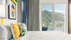 Ein Hotelzimmer mit Bett, Blick auf eine gebirgige Landschaft durch eine Schiebetür und einem gerahmten abstrakten Gemälde an der Wand.