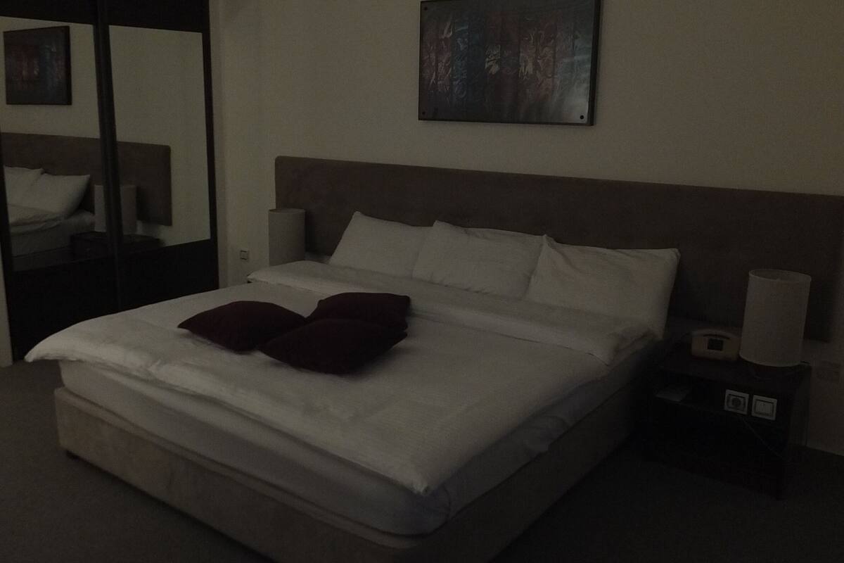 A dimly lit hotel room with a large bed, a nightstand, and a painting on the wall.