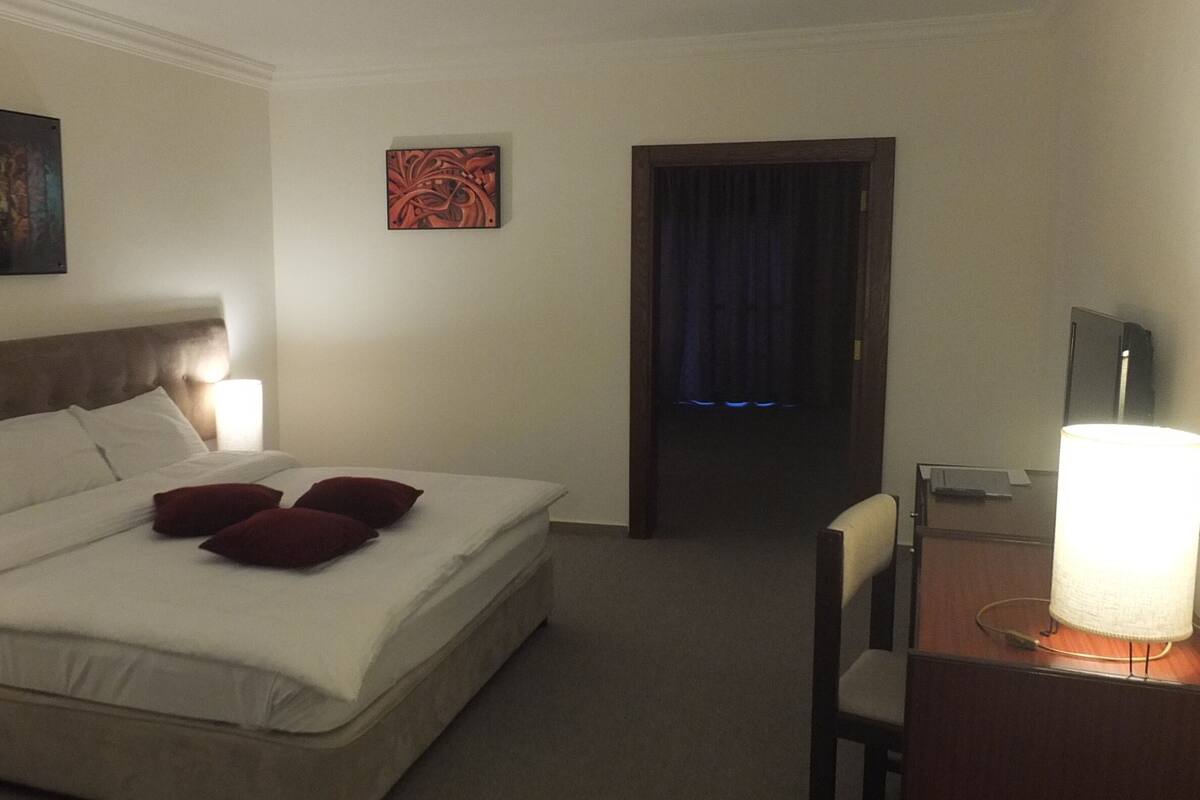 A hotel room with a bed, a desk, a chair, a lamp, and artwork on the walls.