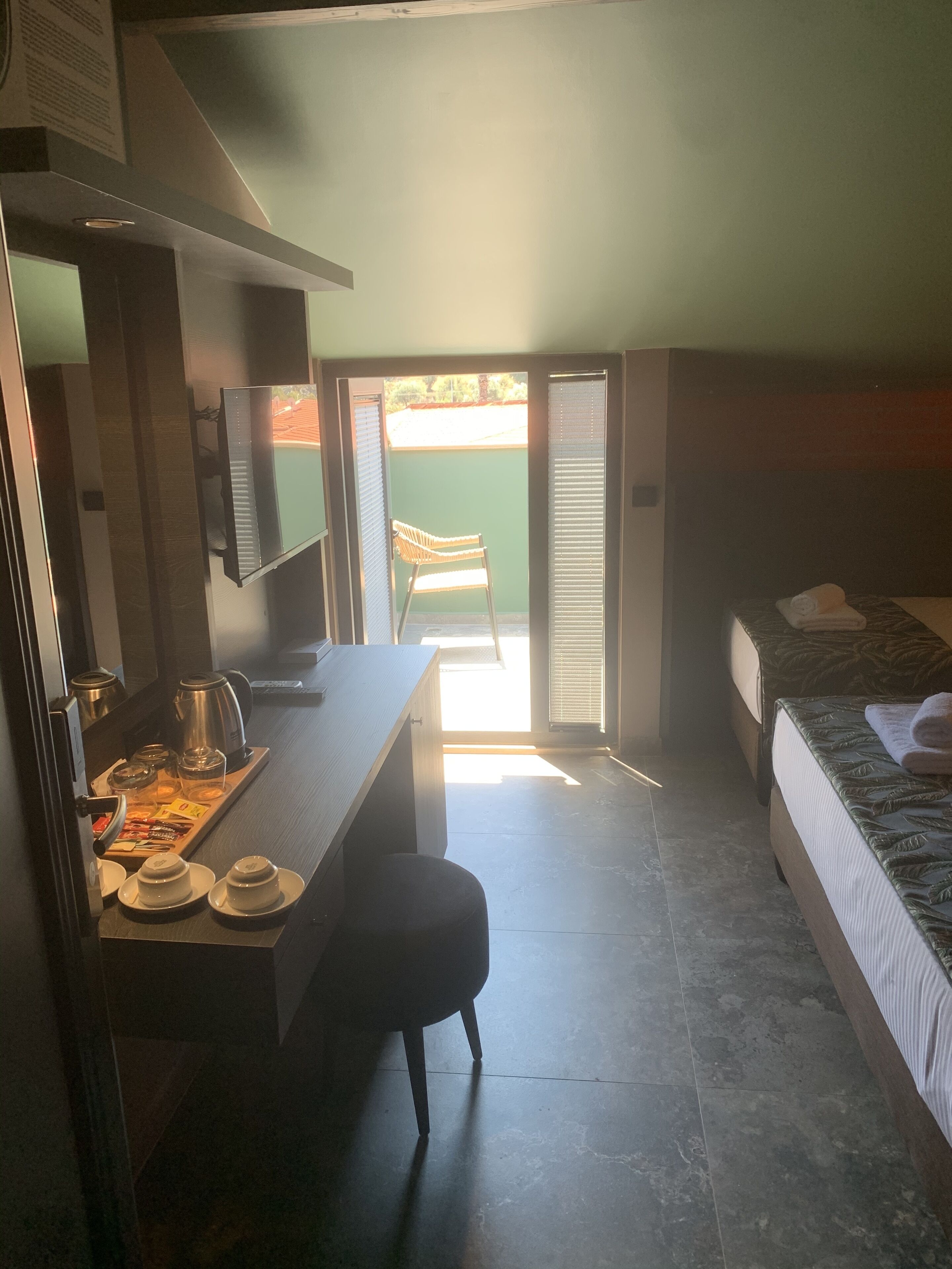 A compact hotel room with a bed, a small desk with a kettle, and a chair.