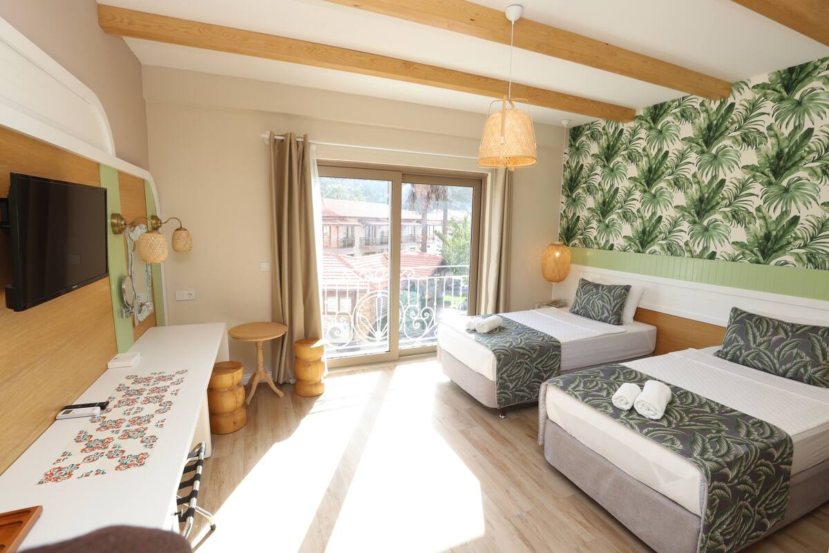 Una habitación de hotel con un ventanal grande, una cama con colcha estampada con hojas, un escritorio con un rompecabezas y un televisor en la pared.