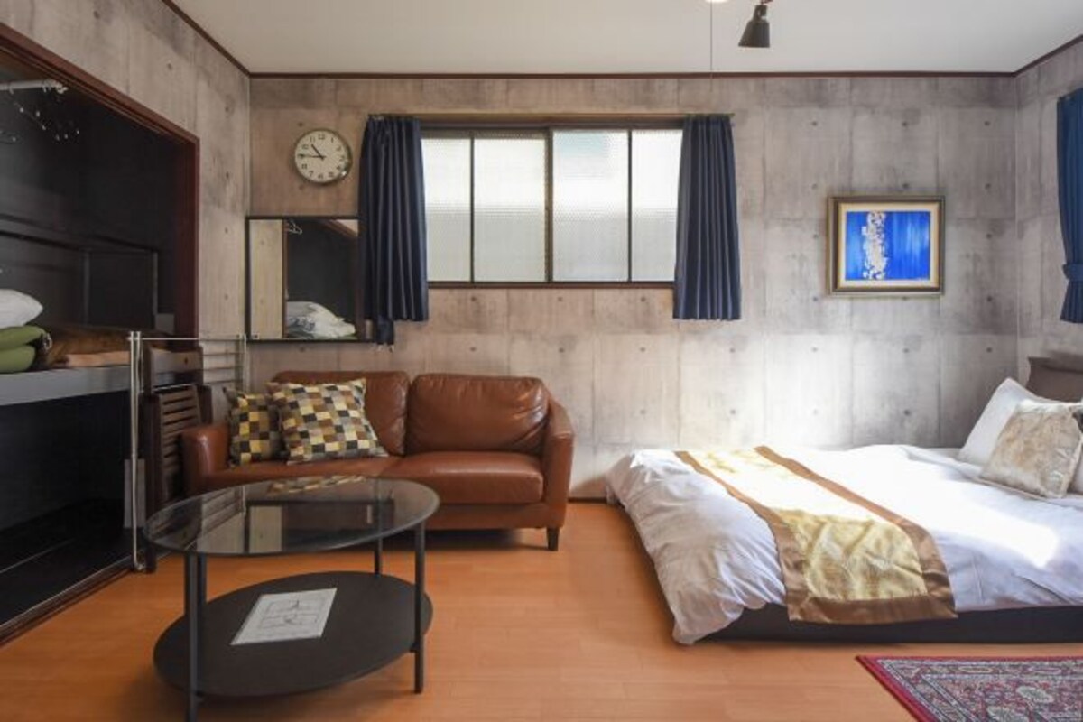 A bedroom with a bed, a brown leather sofa, a glass coffee table, a wooden floor, a window with curtains, a clock, and a framed picture on the wall.