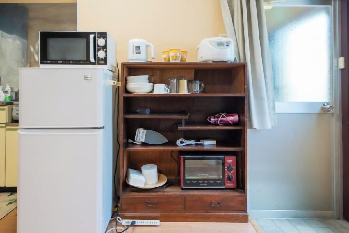 A compact kitchen with a white refrigerator, a microwave, a toaster, and a wooden shelf unit with various kitchen items.