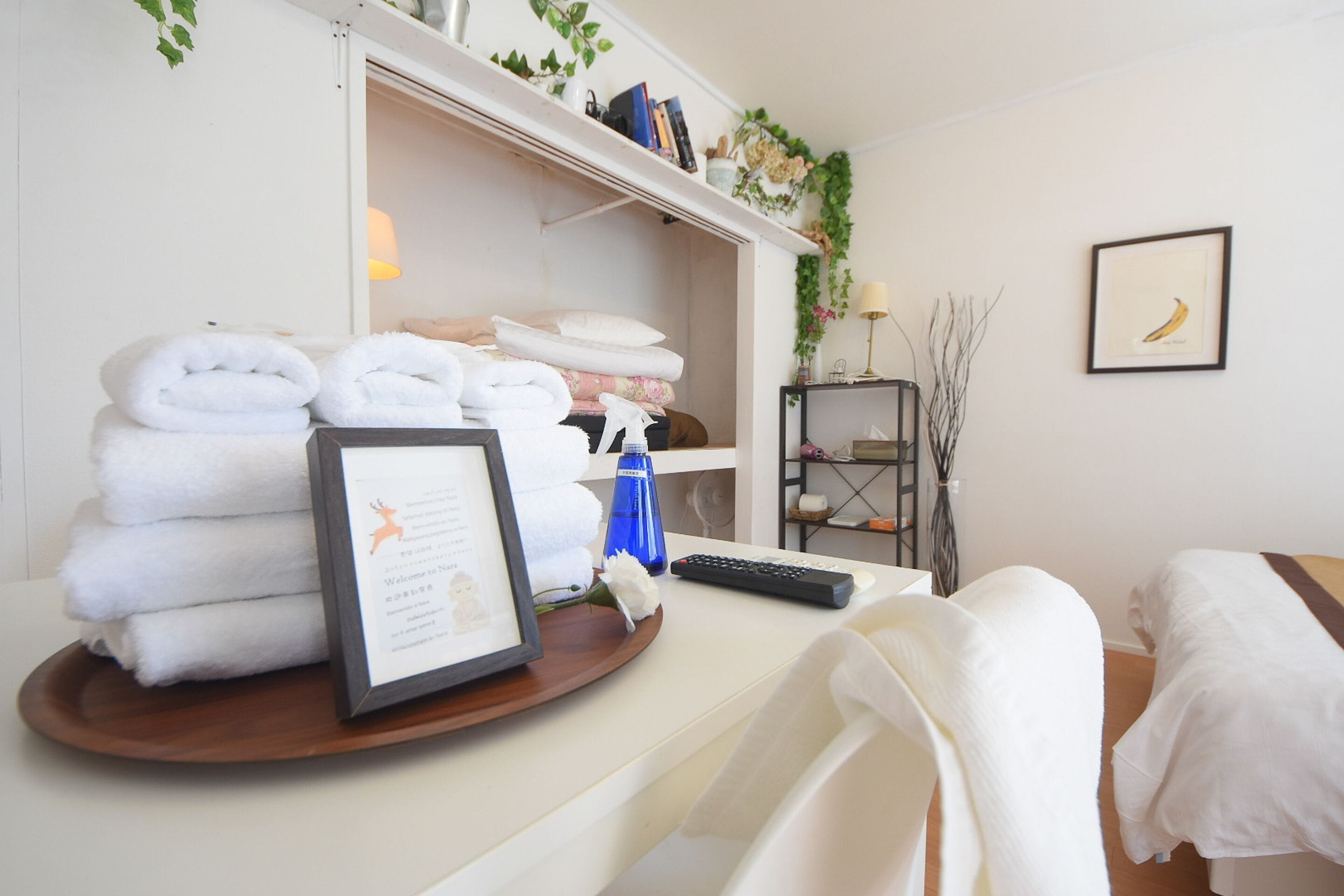 Ein Hotelzimmer mit einem ordentlich bezogenen Bett, einem Schreibtisch mit einer gerahmten Willkommenskarte, einem Stapel Handtücher und einem Regal mit Büchern und Dekoration.