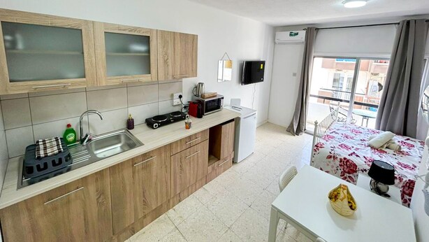 Quarto de hotel compacto com cozinha compacta, cama com colcha florida, mesa pequena para refeições e janela com cortinas.