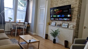 Una sala de estar con una televisión montada en una pared de ladrillo, una zona de comedor con una mesa y sillas, y un sofá.