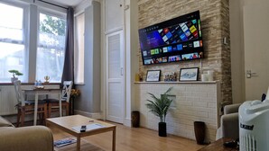 Una sala de estar con un televisor de pantalla plana montado en una pared de ladrillo, una chimenea, una zona de comedor con una mesa y sillas, y una planta en maceta.
