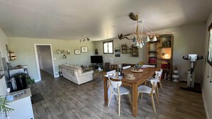 Un'ampia zona giorno con un tavolo da pranzo in legno, sedie bianche, un divano e un televisore.