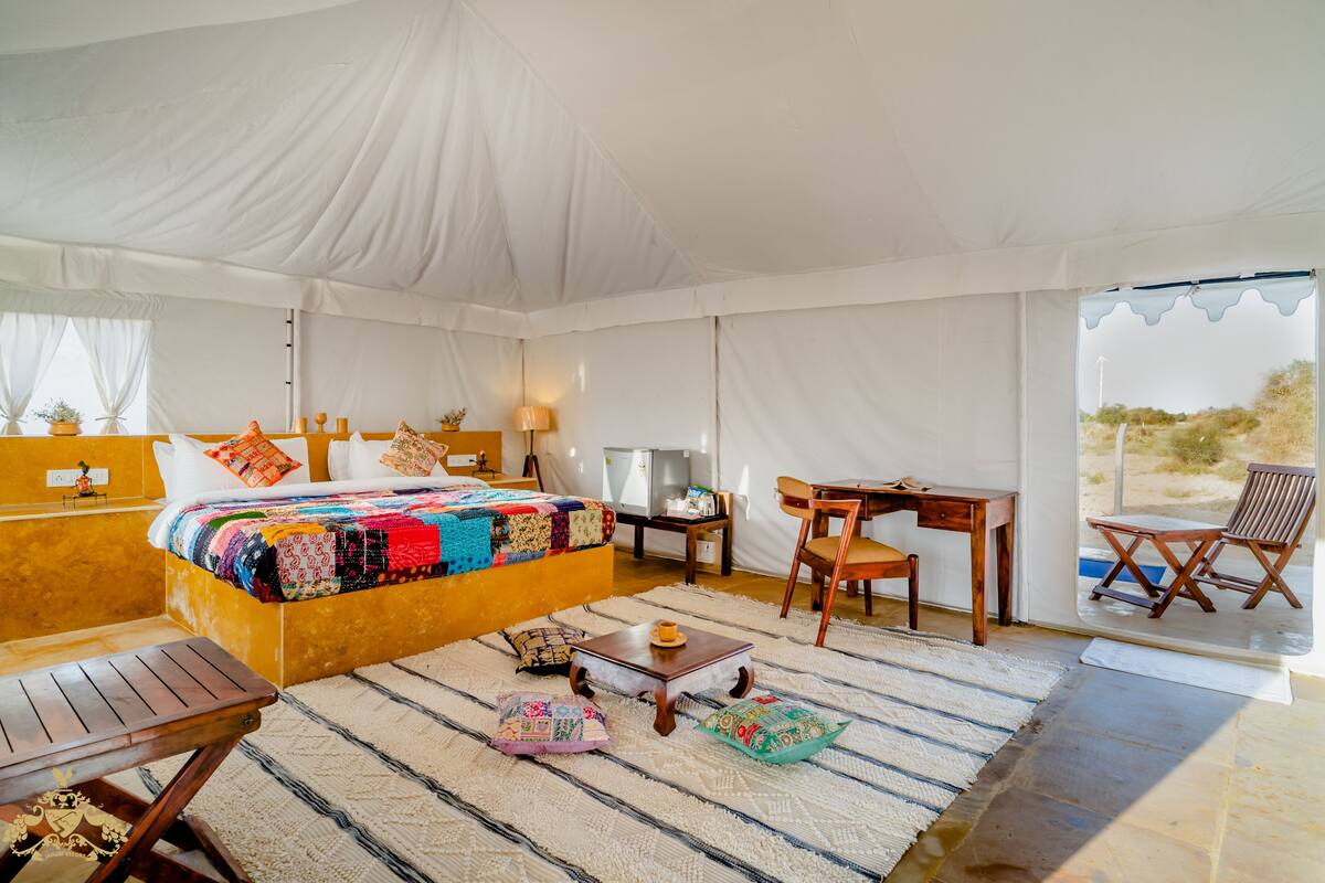 Alojamiento tipo tienda con cama, escritorio, sillas y vistas al exterior.