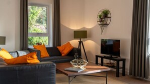 Un salon moderne avec un canapé en cuir noir, une table basse en bois, un lampadaire, un téléviseur posé sur un meuble et une fenêtre avec des rideaux.
