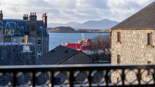 A coastal view with stone buildings, a red-roofed structure, and a distant mountain range.
