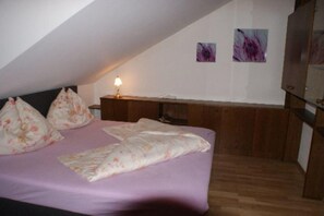 Une chambre avec un plafond en pente, un lit recouvert d’une literie à motifs floraux, une table de chevet avec une lampe, et deux œuvres d’art abstraites accrochées au mur.