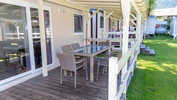 A covered outdoor area with a glass table and wicker chairs, wooden deck, and a view of a grassy yard.