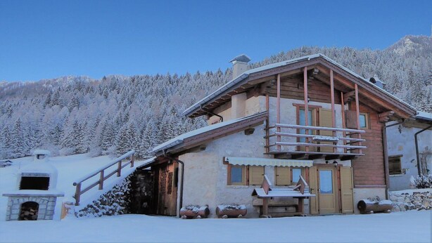 Una cabaña de madera con balcón, chimenea de piedra y un bosque nevado al fondo.
