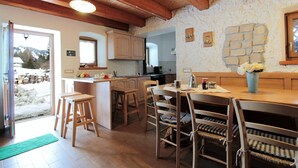 Una cocina rústica con vigas de madera, pared de piedra y zona de comedor con sillas y mesa de madera.