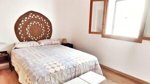 Ein Schlafzimmer mit Bett, Nachttisch, einer Nachttischlampe, einem gewebten Kopfteil, einem Fenster mit Holzrahmen und einer kleinen weißen Bank.