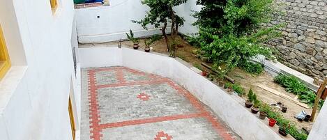 Un patio con zona pavimentada que presenta diseños geométricos, rodeado por un edificio blanco con muro de piedra y un árbol.