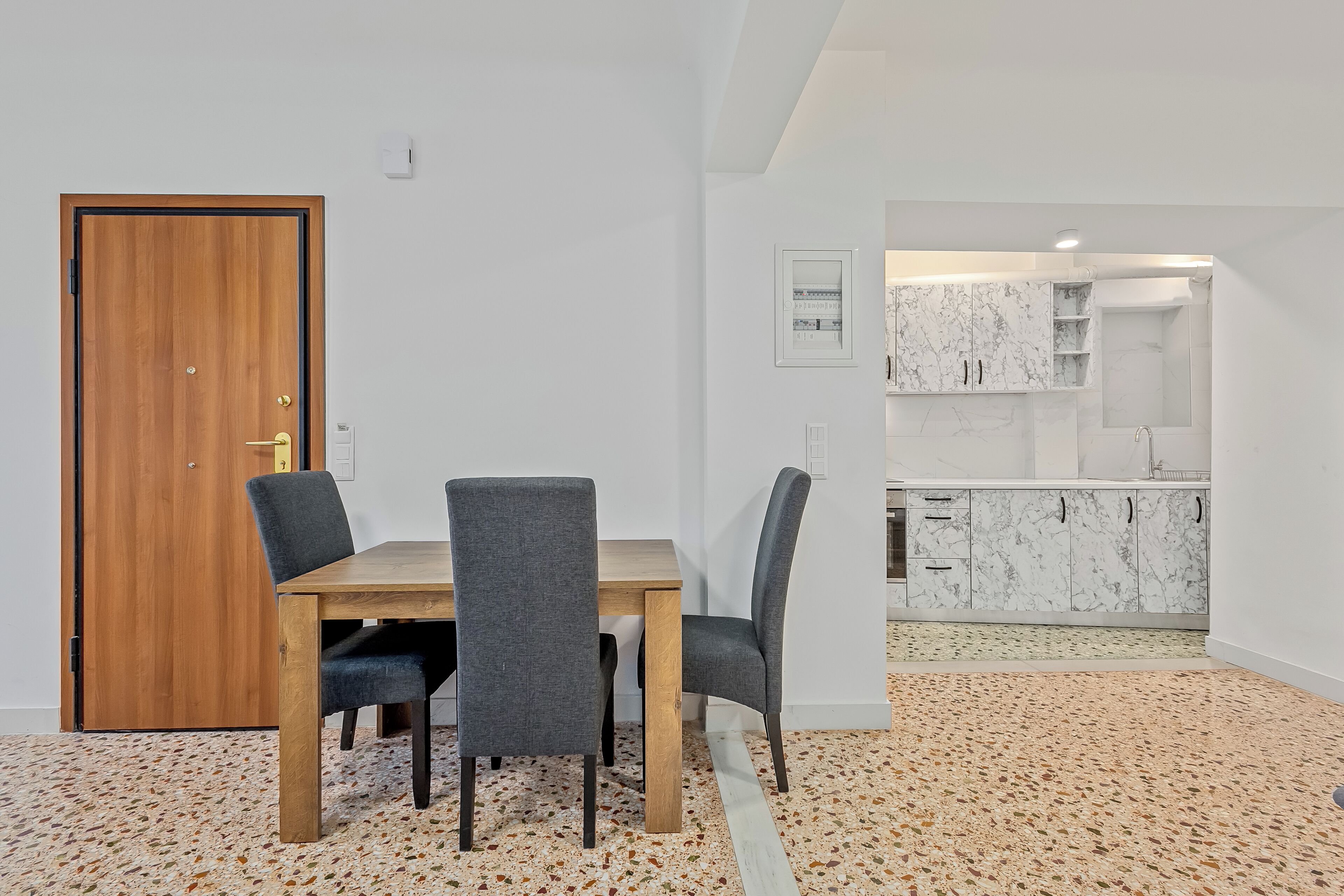 Un comedor con una mesa de madera y cuatro sillas, una cocina con muebles blancos de mármol y un suelo de terrazo.