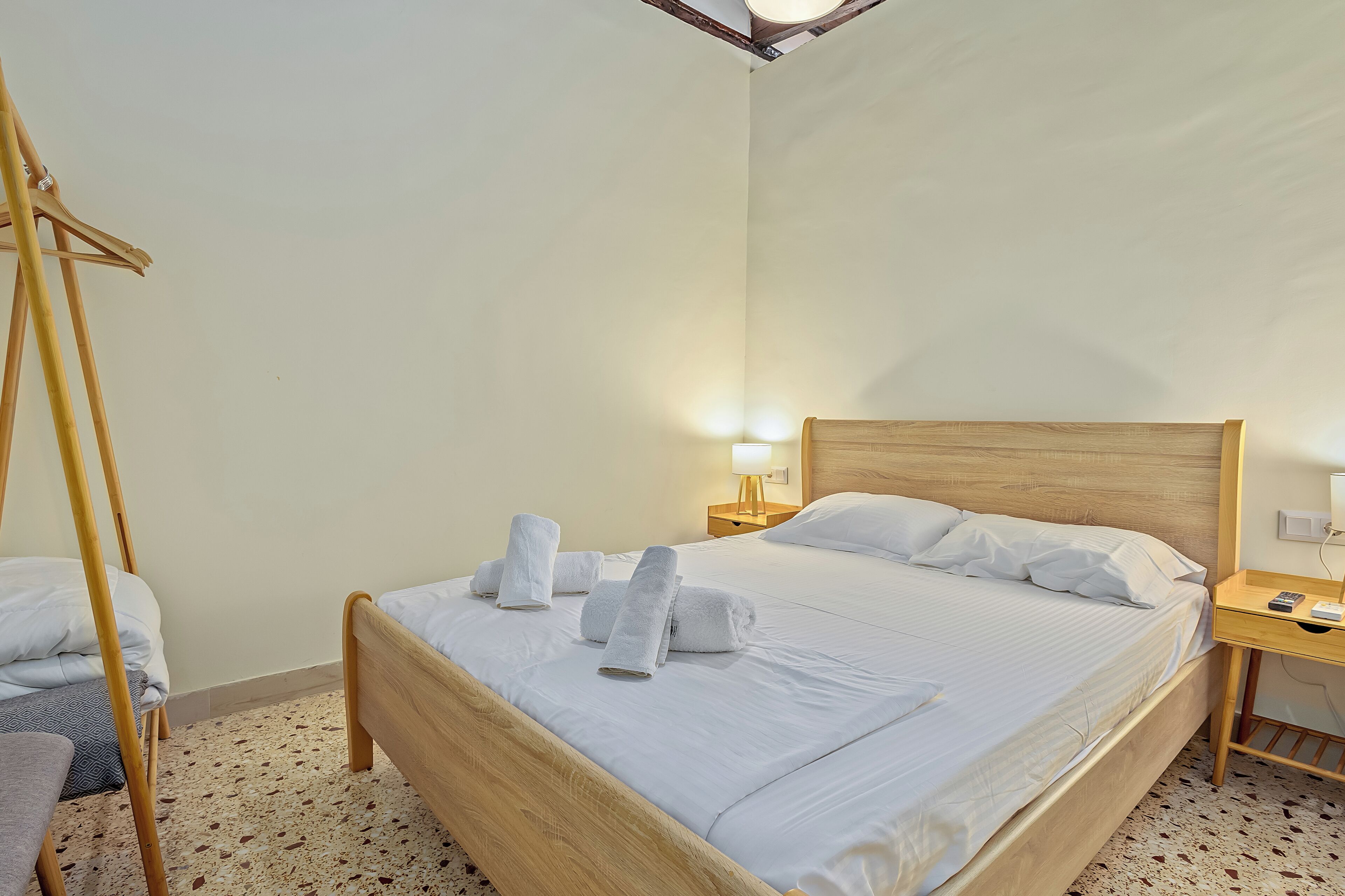 A hotel room with a wooden bed, white linens, a wooden nightstand with a lamp, and a wooden chair.