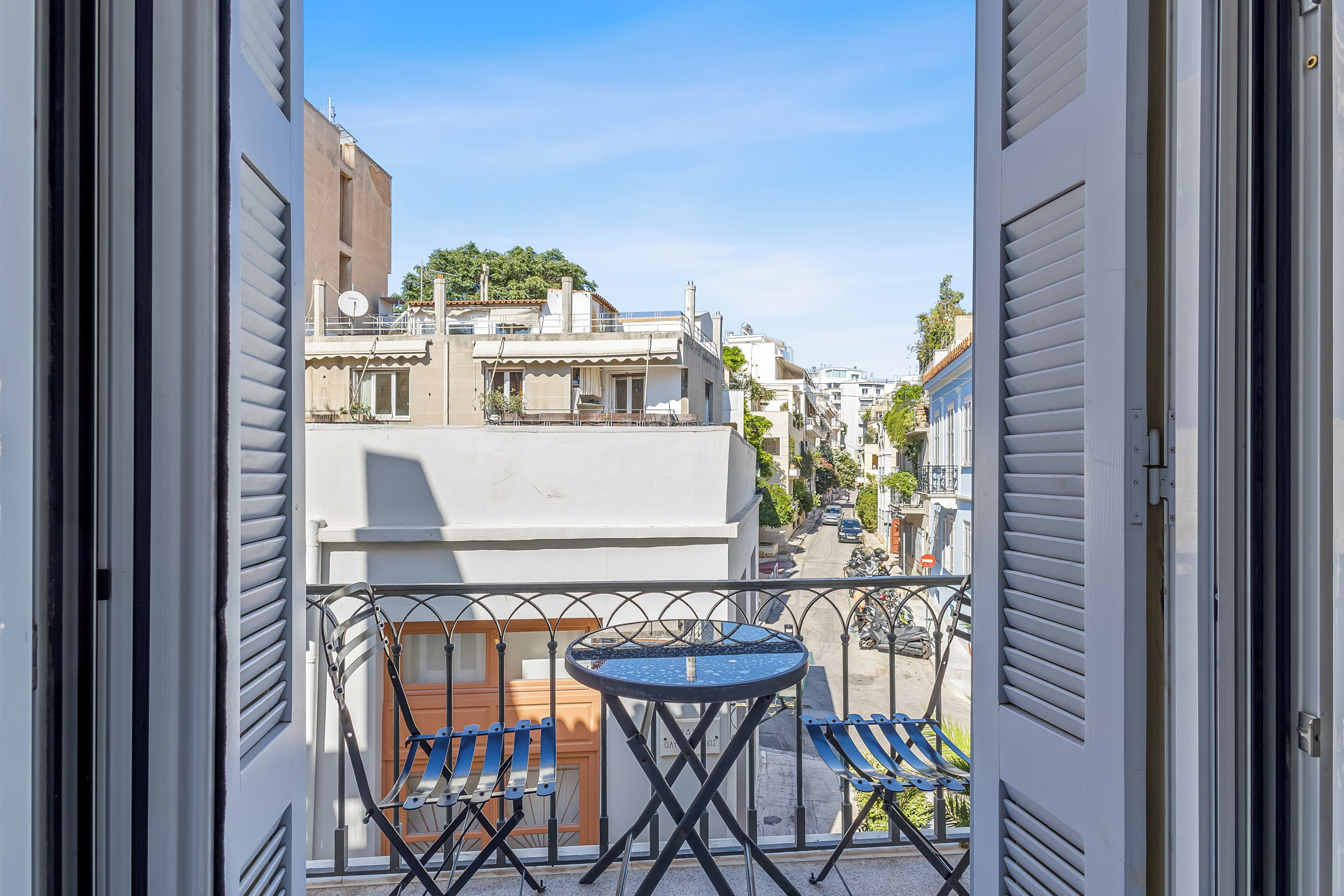 Un balcón con una mesa y dos sillas, con vistas a una calle con edificios y un cielo despejado.