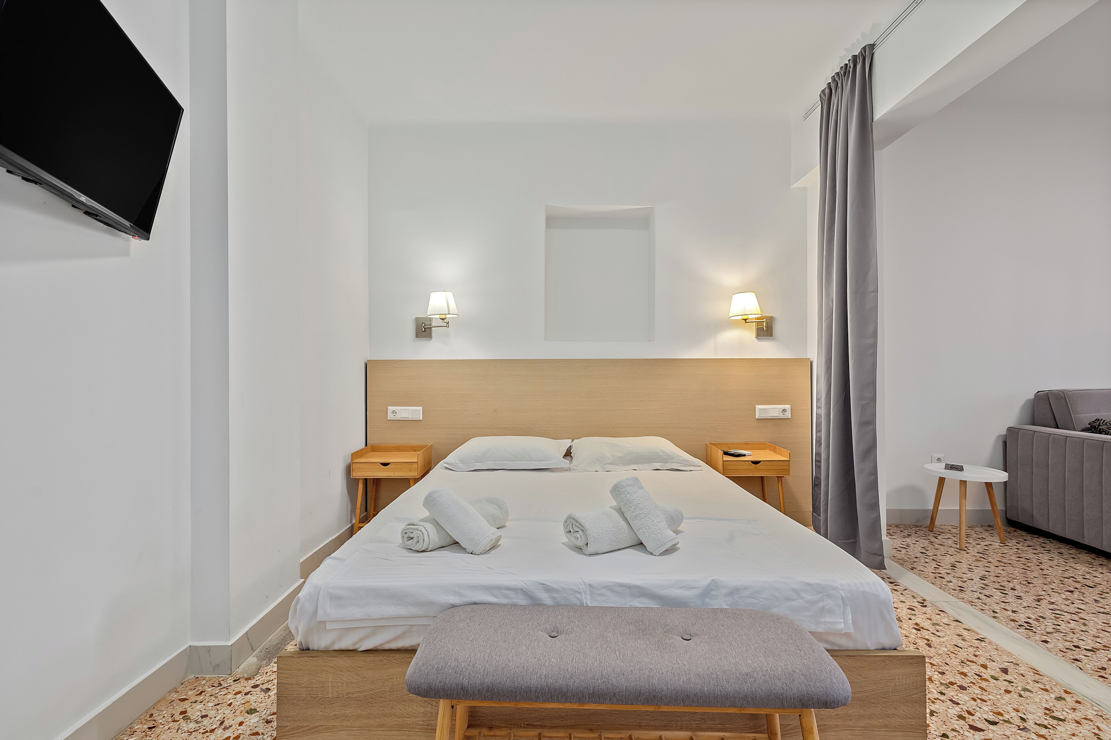 A hotel room with a bed, bedside tables, a TV mounted on the wall, and a grey sofa.