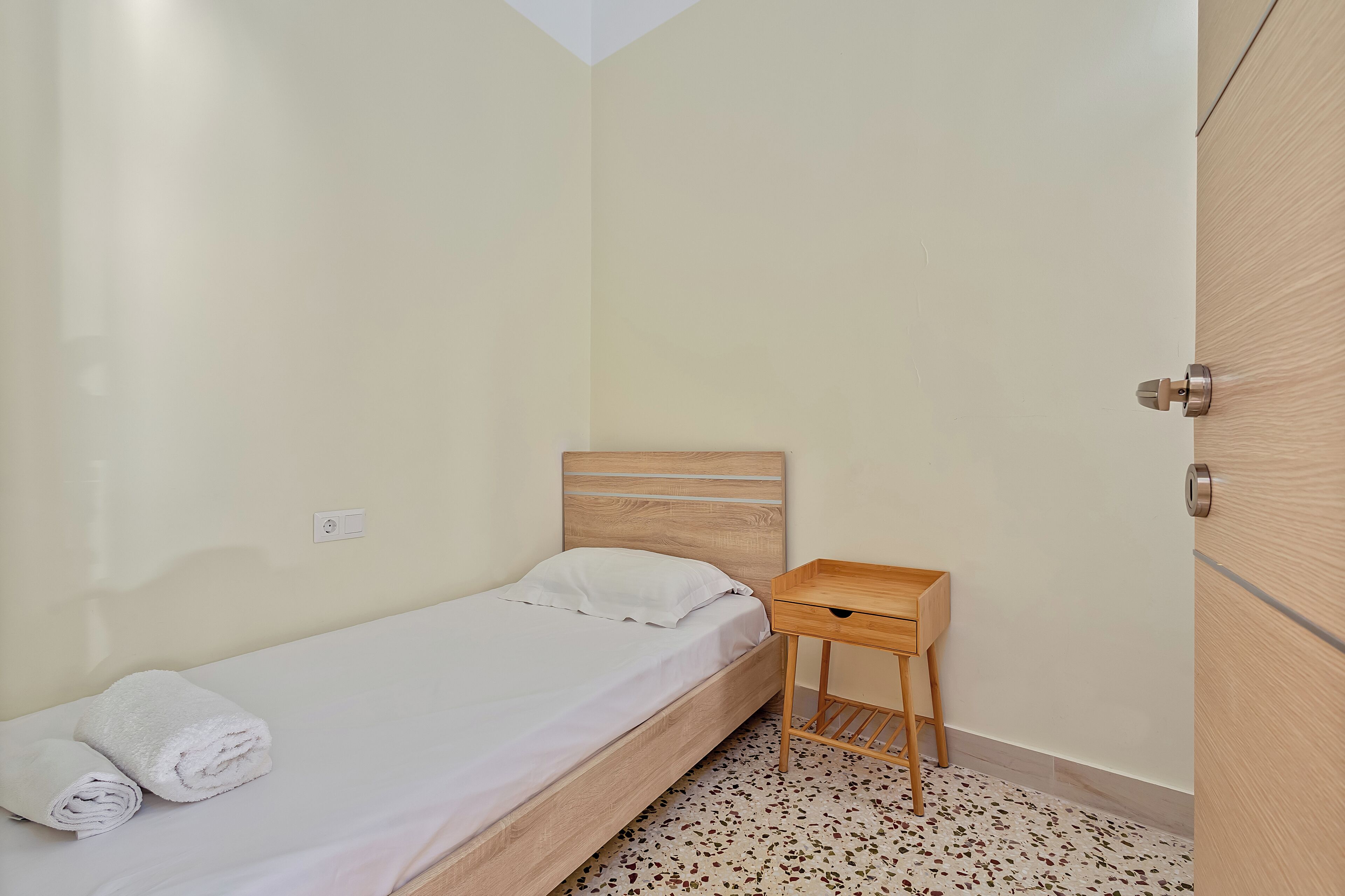 Una habitación pequeña con una cama individual, cabecera de madera, mesita de noche de madera con un cajón y una puerta con tirador metálico.