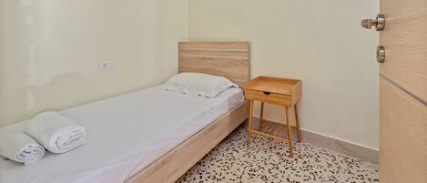 Una habitación pequeña con una cama individual, cabecera de madera, mesita de noche de madera con un cajón y una puerta con tirador metálico.