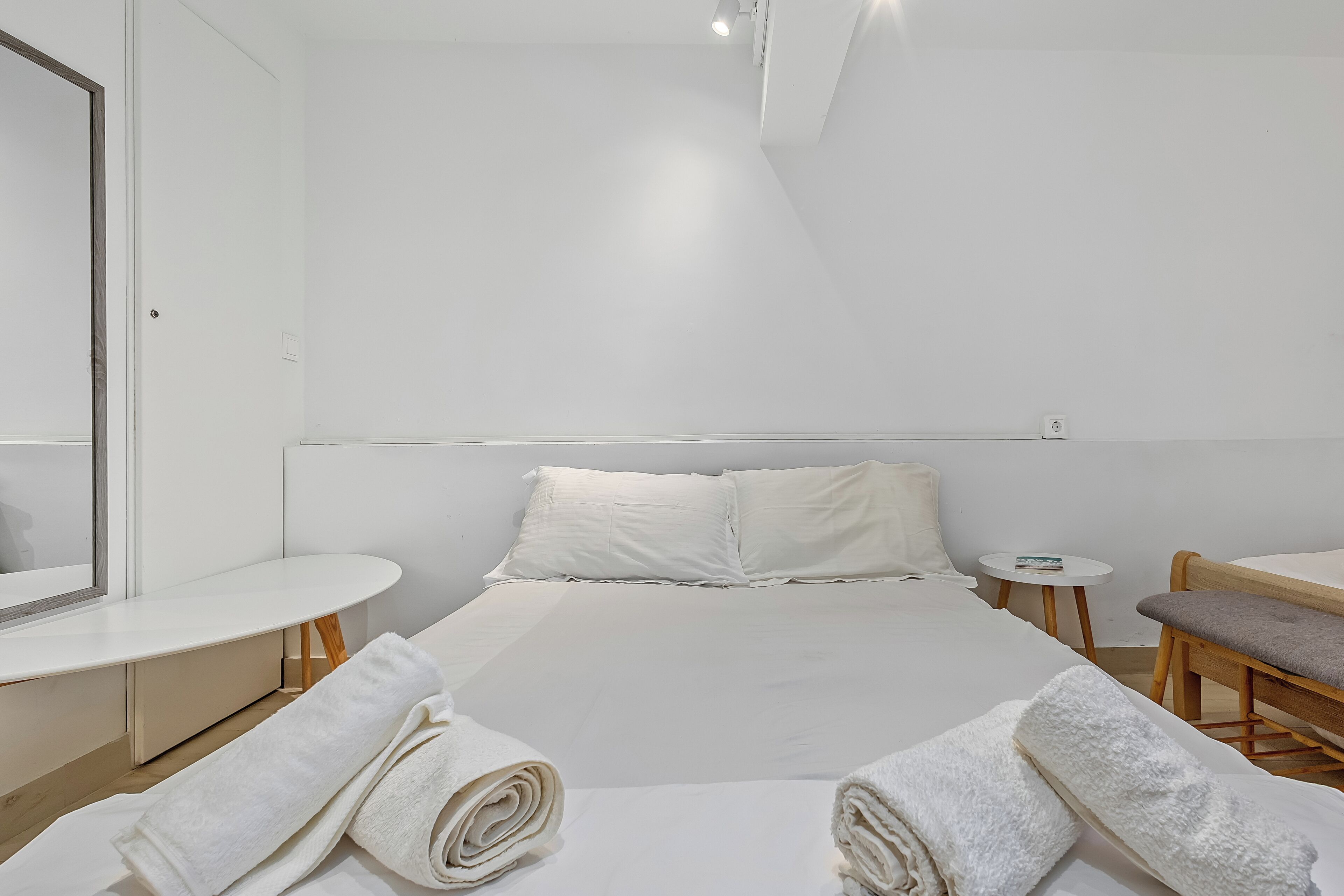 Habitación de hotel minimalista con una cama, una mesita redonda, una mesita de noche pequeña, una silla y un espejo.