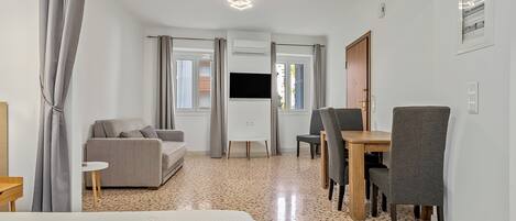A hotel room with a bed, a sofa, a dining table with chairs, a TV, and a window with curtains.