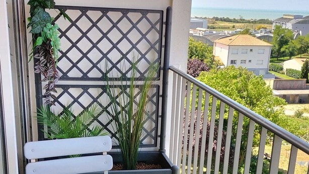 Un balcone con un tavolo bianco, due sedie bianche, una piccola pianta in vaso e vista sul mare e sugli edifici.