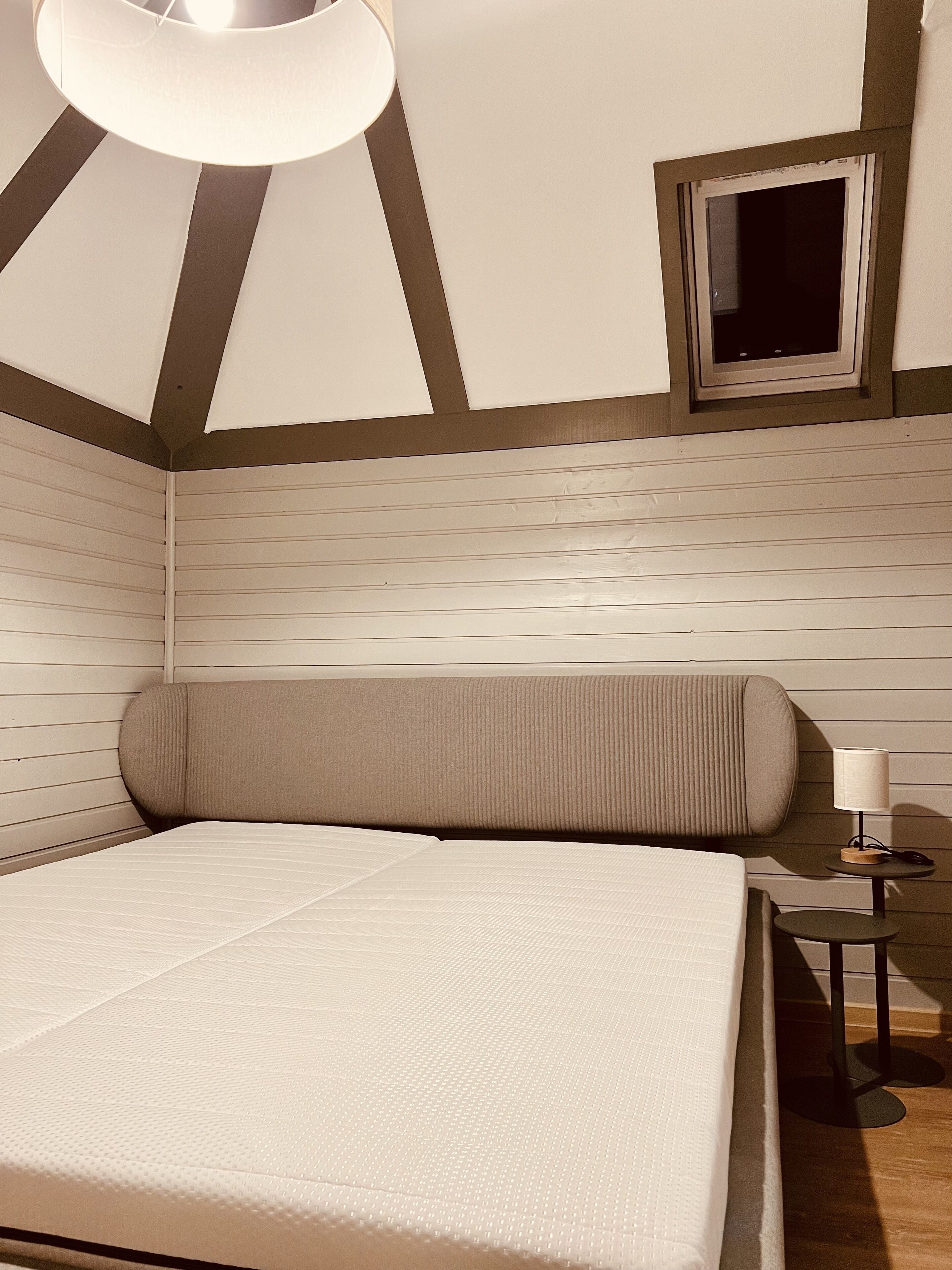 A bedroom with a large bed, a small table with a lamp, and a TV on the wall.