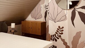 Une chambre à coucher avec un lit, une commode en bois, un miroir rond et un plafond avec des poutres apparentes.