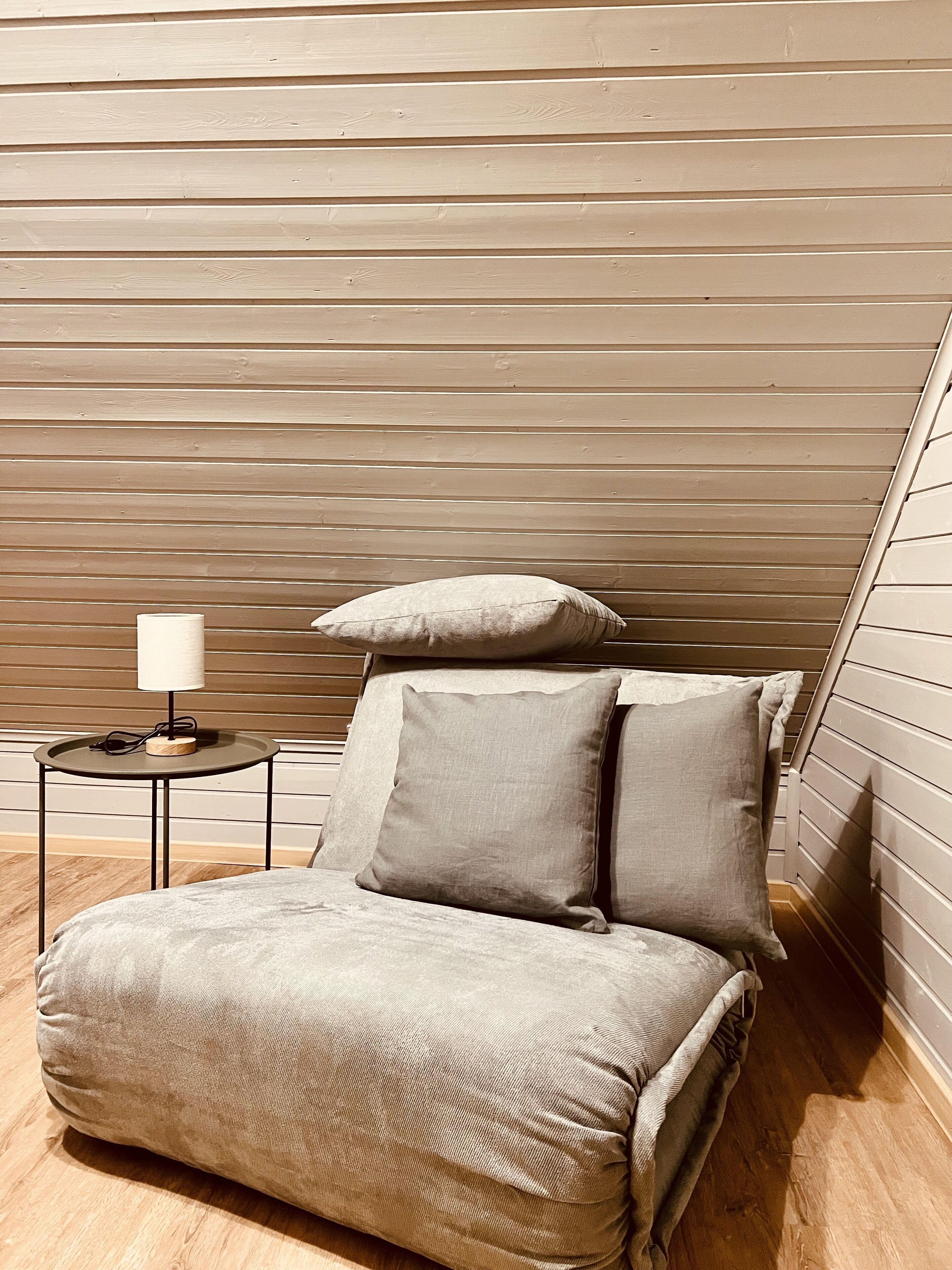 Un dormitorio pequeño y acogedor con techo inclinado, una cama individual con dos almohadas, una mesita de noche con una lámpara y una mesa redonda con un ordenador portátil.