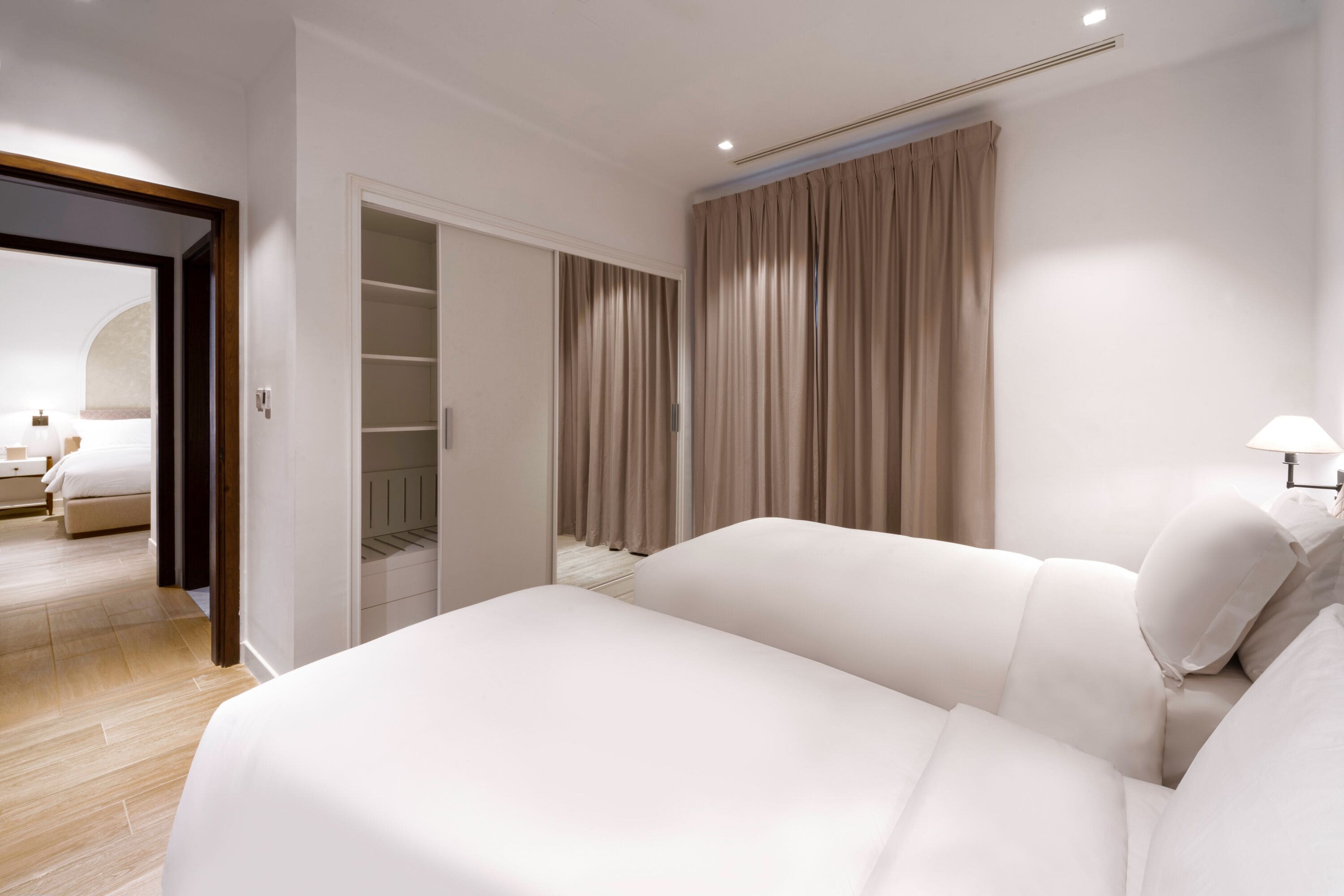 A modern hotel room with a large bed, a walk-in closet, and a view of the adjacent room.