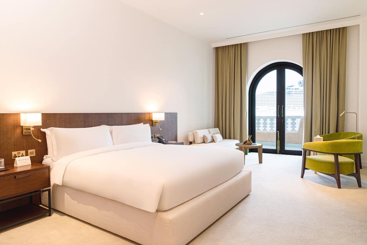 A hotel room with a large bed, a nightstand, a desk, a chair, and a view of the outside through a window.